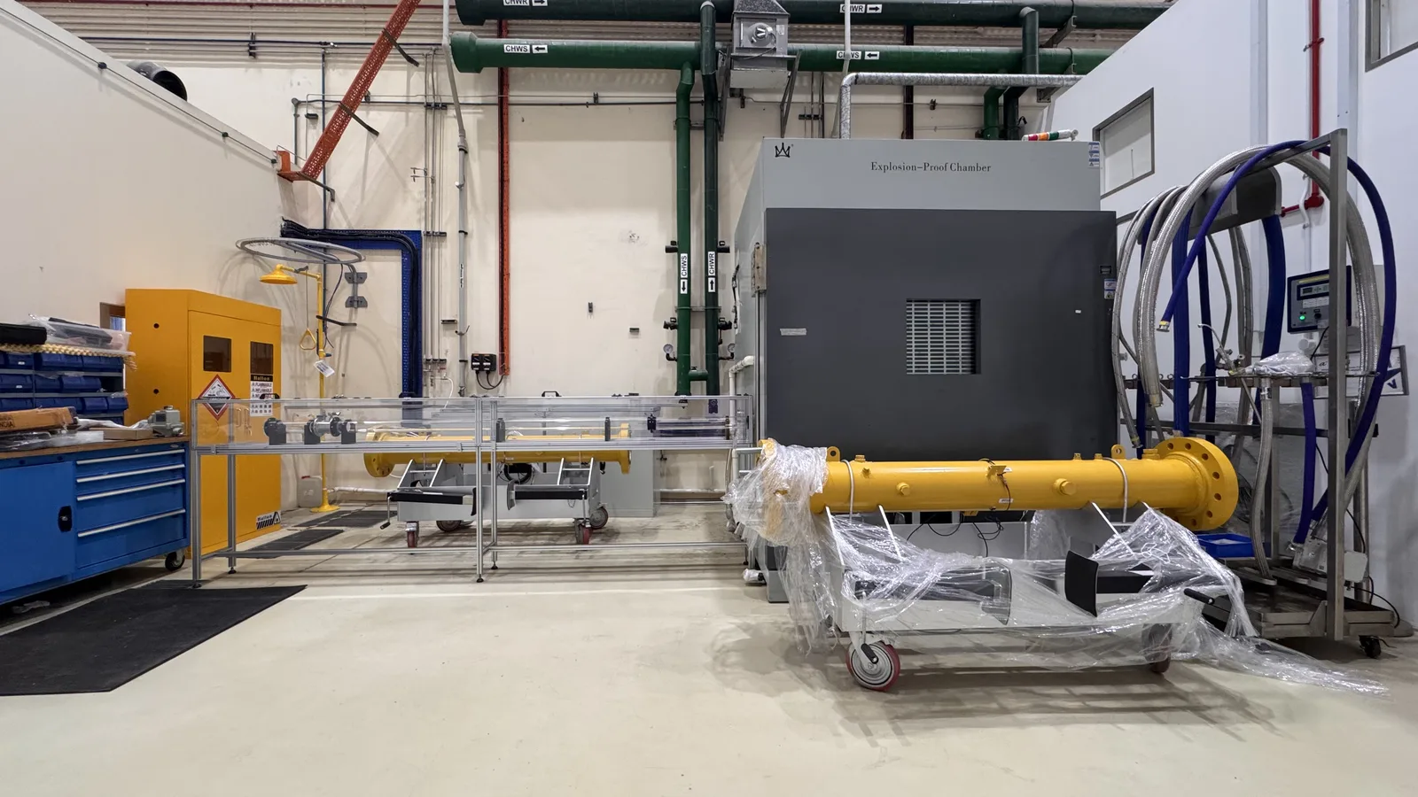 The lab from the floor: the mini detonation tube in yellow on its wrapped trolley in front of the grey explosion-proof chamber, with the full tube running behind its acrylic guarding on the left.