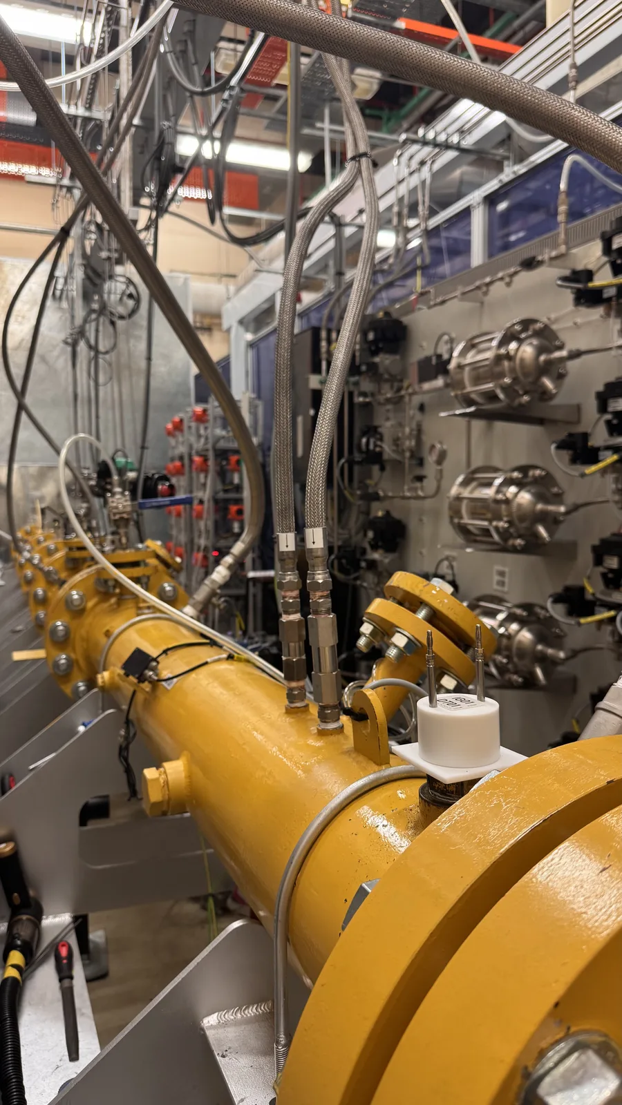 Looking down the yellow detonation tube from beside a sensor station: an igniter cap and braided sensor lines on top of the spool, flanged joints marching away, the gas panel's regulators on the wall to the right.
